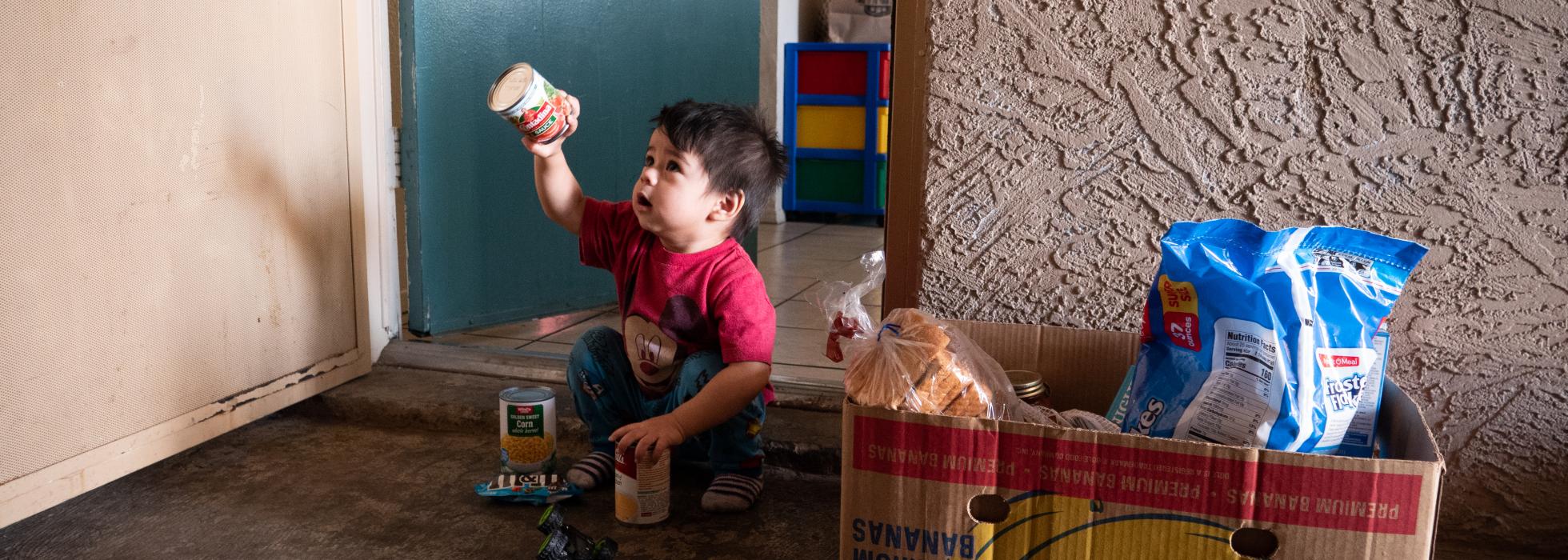 Young boy sitting outside his home with a food box holding up a food item
