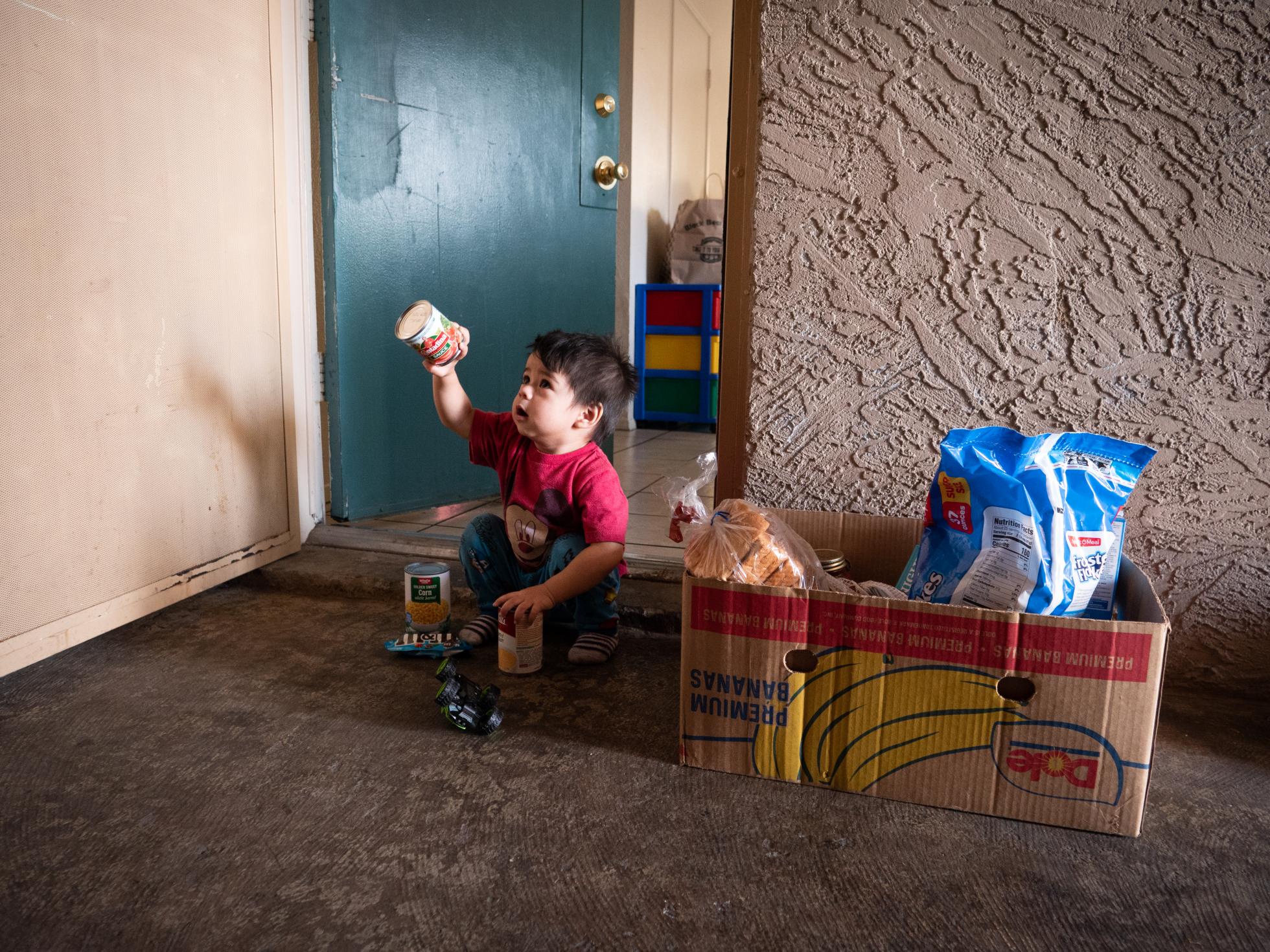 Young boy sitting outside his home with a food box holding up a food item