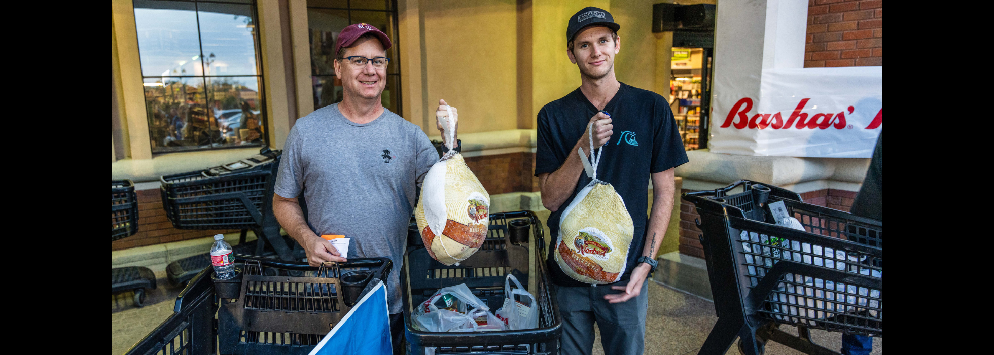 turkey-tuesday-content-hub-header Two men standing in front of a Bashas' store holding up frozen turkeys